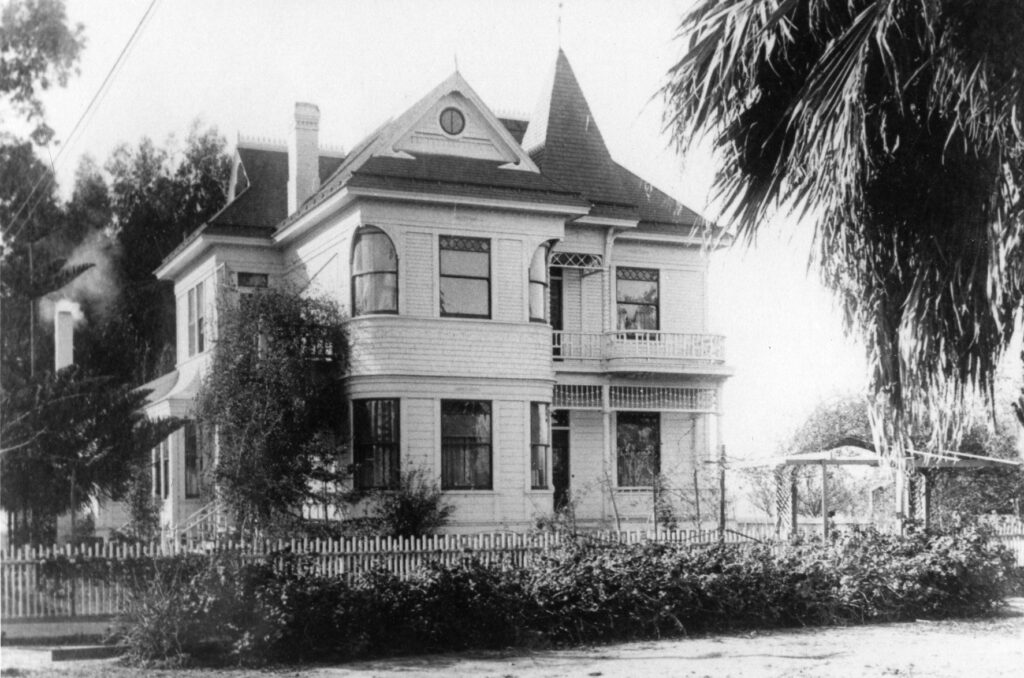 Black-and-white historical photograph of the two-story Victorian Justin Petit Ranch House, featuring a turret, arched windows, balcony, and picket fence, captured before relocation to Heritage Square.