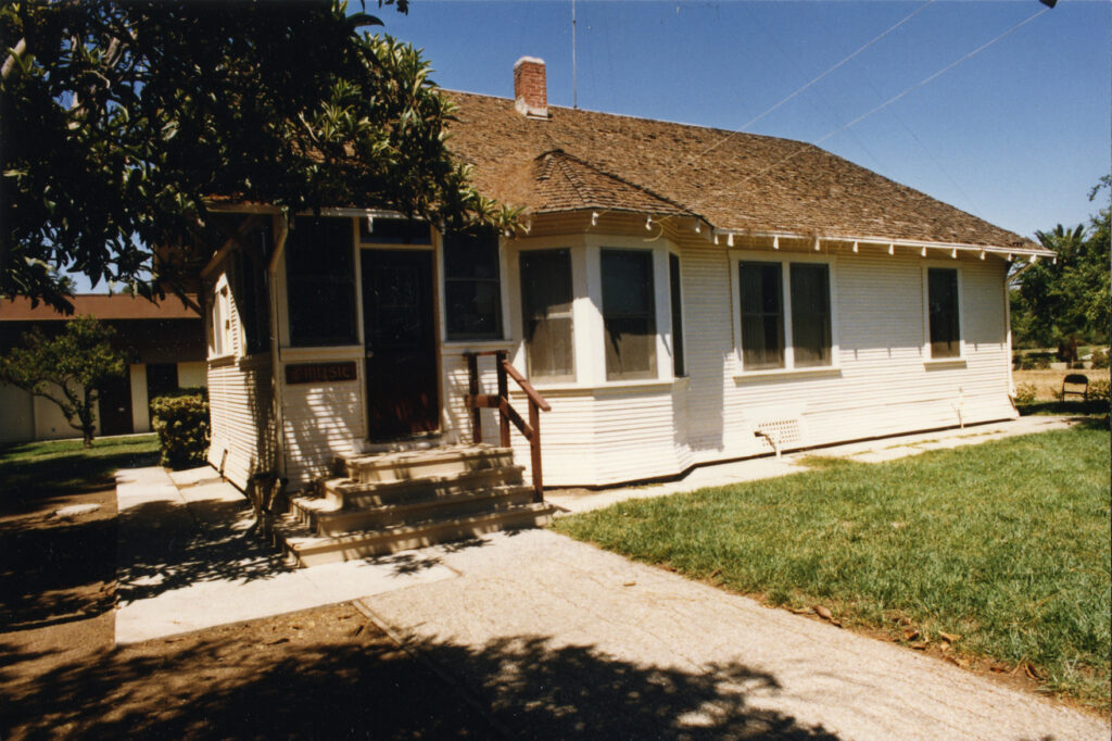 White single-story house with shingled roof, porch steps, and a "MUSIC" sign on the door, surrounded by trees and lawn, circa 1980s.