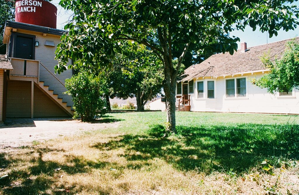 A ranch property features a red "PEDERSON RANCH" water tower atop a beige building, a white house with a brown roof, and a large tree on a grassy lawn.