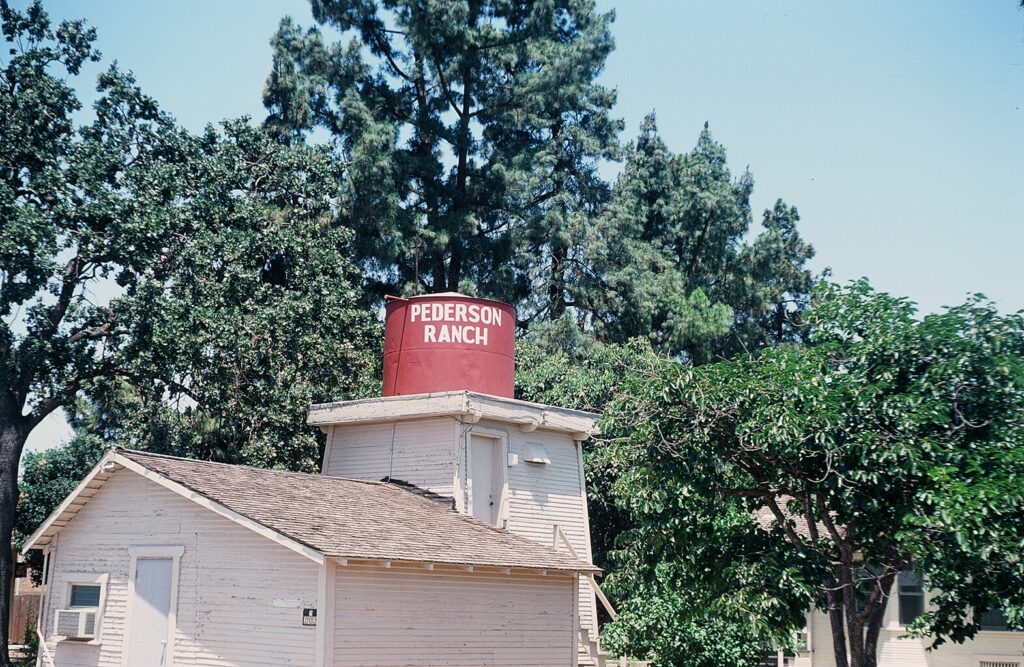 A white wooden building features a red water tower labeled "PEDERSON RANCH" surrounded by lush green trees under a clear blue sky.