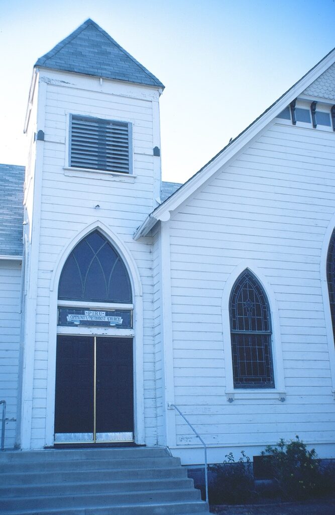 White wooden church with steeple, arched windows, and "PIRU COMMUNITY METHODIST CHURCH" sign above double doors. 1981 photograph.