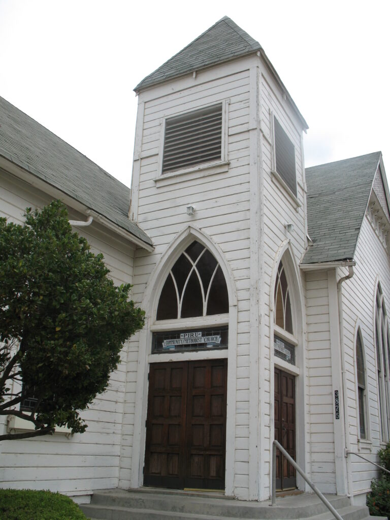 White wooden church with Gothic arched windows, double doors, and a sign reading "Piru Community Methodist Church" with address number 3875.