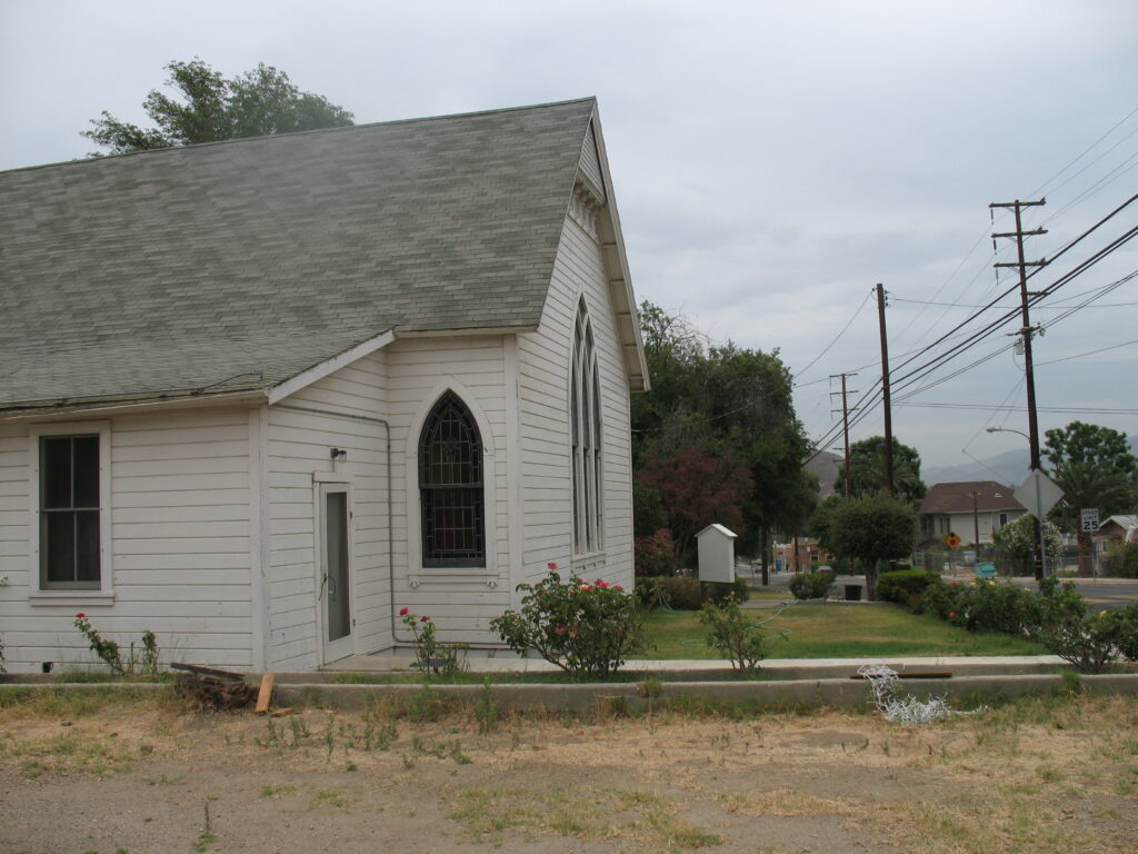 White clapboard Piru Methodist Church with gray shingled roof, Gothic arched windows, and a small garden with red flowers. Utility poles and a 25 mph speed limit sign are visible in the background.