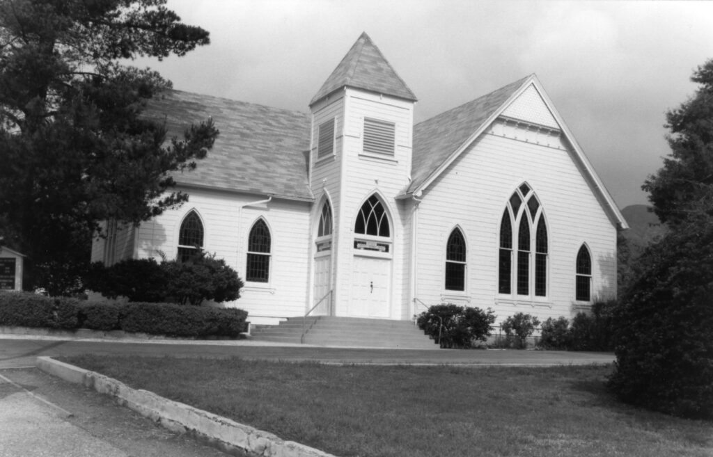 White clapboard church with Gothic arched windows, central entrance steps, and surrounding trees and shrubs, circa 1980s.