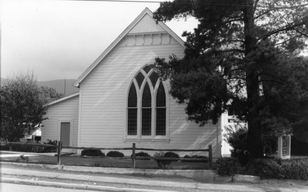 White wooden church with gable roof and large stained-glass window, surrounded by trees and a wooden fence, circa 1980s.