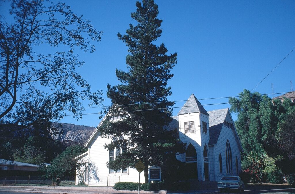 White Piru Methodist Church with steeple and arched windows, surrounded by trees and a yellow "Not a Through Street" sign, with mountains in the background, photographed in 1981.
