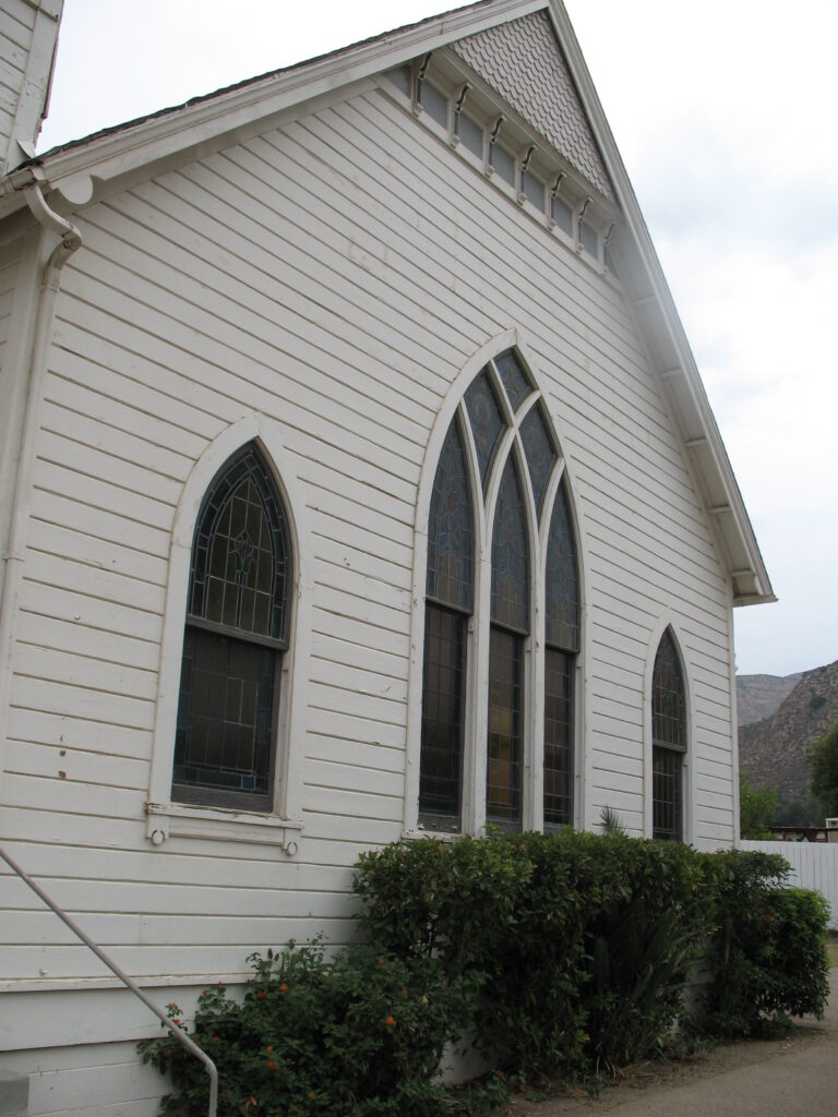 White clapboard Piru Methodist Church exterior features arched stained glass windows with leaded glass panes, a gable roof with decorative trim, and green shrubs at the base.