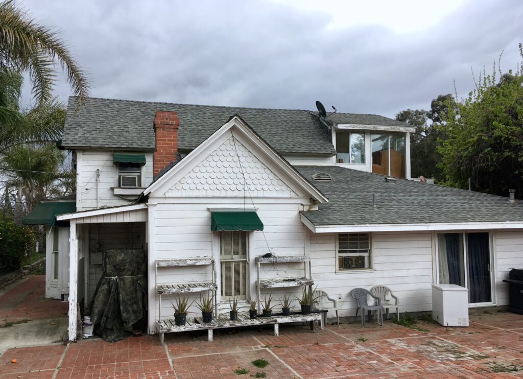 White two-story house with green awnings, brick patio, wooden plant shelves, covered carport, and satellite dish; captioned 'Tanner Homestead, 2019. Credit: Craig Baker'