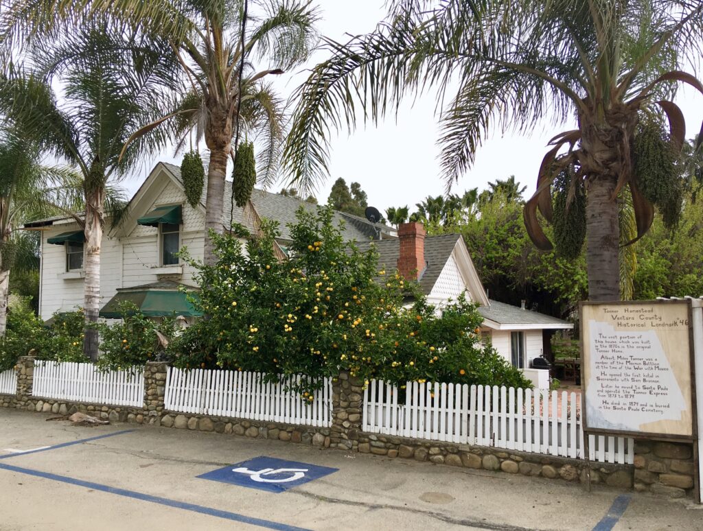 Tanner Homestead, a white house with green awnings, surrounded by palm trees and citrus trees. Features a white picket fence, stone base, historical marker sign with text, and blue handicap parking symbol.
