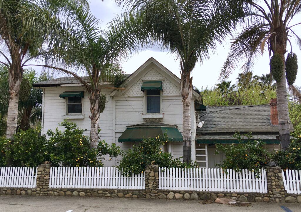 White house with green awnings surrounded by palm trees and citrus trees bearing yellow fruit, enclosed by a white picket fence with stone posts.
