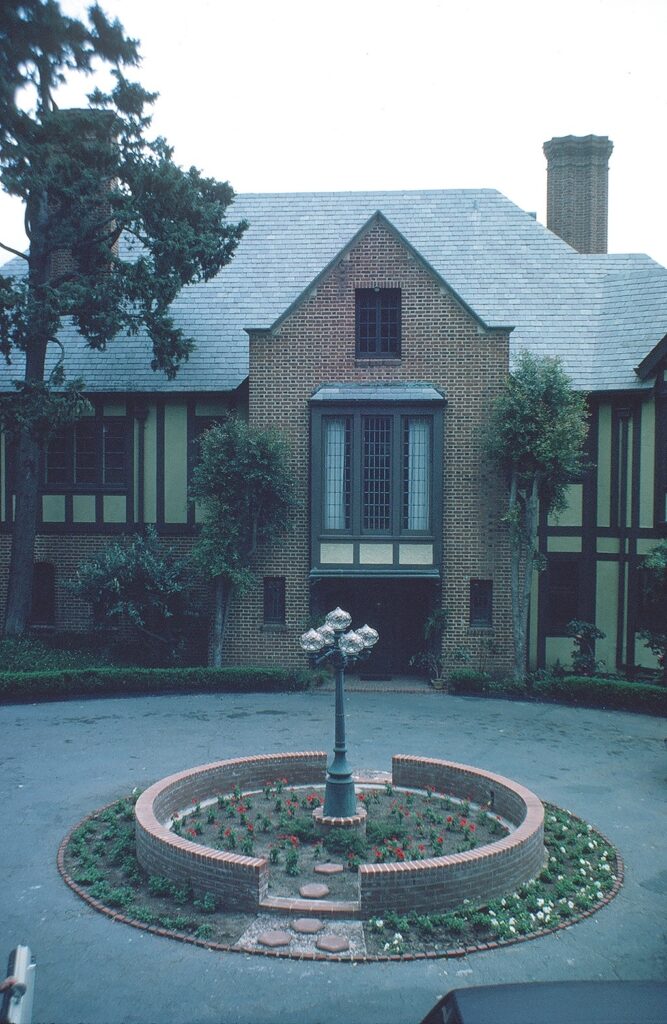 Teague Mansion (1982) features a brick facade with a gray shingle roof, central bay window, and a circular brick garden with flowers and a lamp post in the foreground.