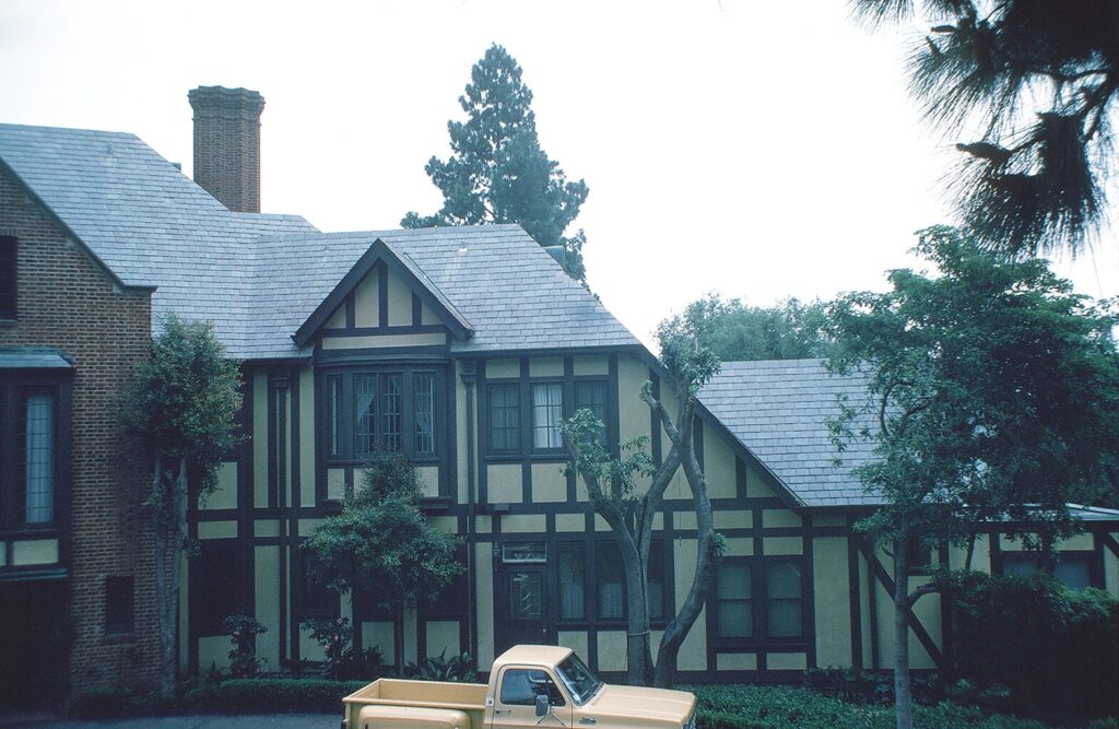 Tudor-style mansion with half-timbered walls, gray shingles, and a brick section, surrounded by trees and a beige pickup truck. Teague Mansion, 1982.