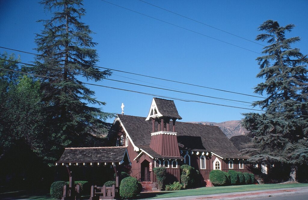 Brown wooden Trinity Episcopal Church with white trim, steeple, and cross, surrounded by trees under a clear blue sky, photographed in 1981.