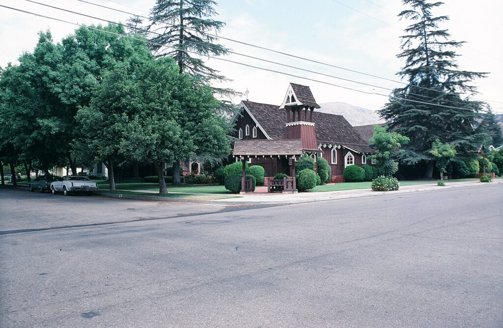 Tudor-style Trinity Episcopal Church with brown shingles and white trim, surrounded by green trees and landscaping, photographed in 1978. Two vintage cars are parked on the street in front of the church.