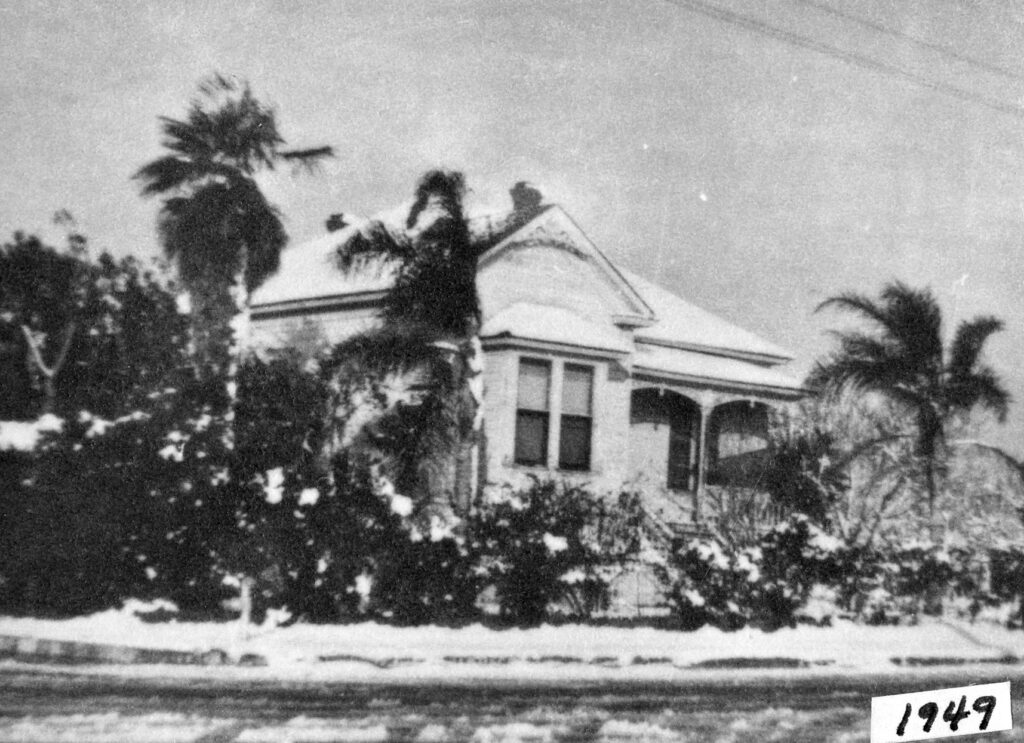 Black-and-white photograph of the Wiltfong House covered in snow, surrounded by palm trees and shrubs, with the year 1949 visible in the bottom right corner.