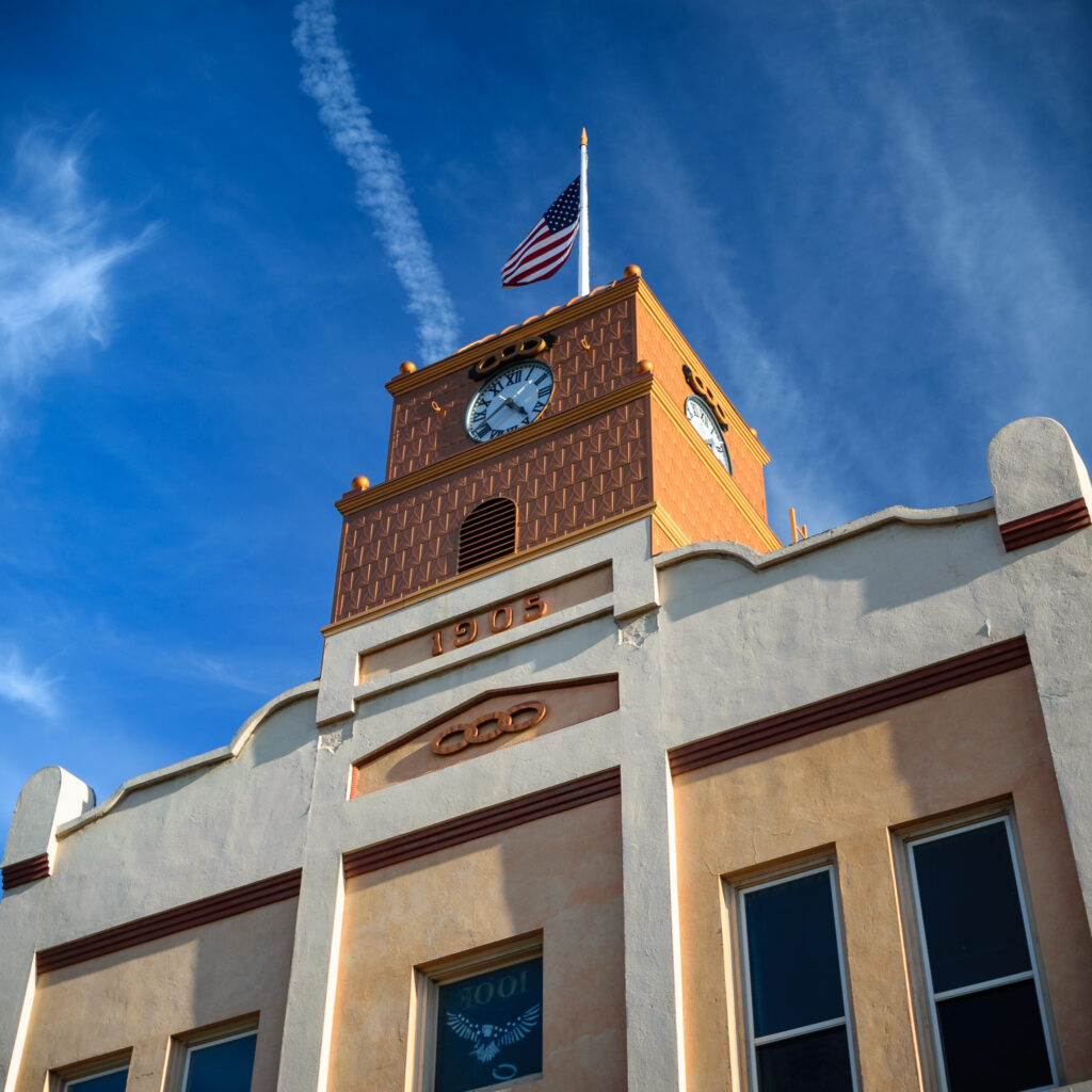 Odd Fellows' Town Clock building with a brown clock tower featuring dual clocks, the year "1905" inscription, an American flag on top, and a beige facade against a clear blue sky.