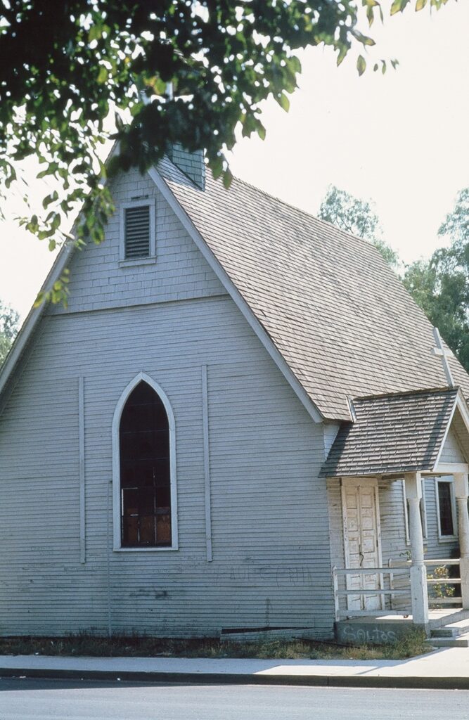 Light gray wooden church with steep roof, arched window, and porch, dated 1978, surrounded by trees.