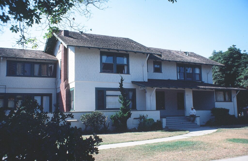 1981 photograph of a two-story white house with a brick chimney, dark shingled roof, and a porch where a person sits, surrounded by trees and a lawn.