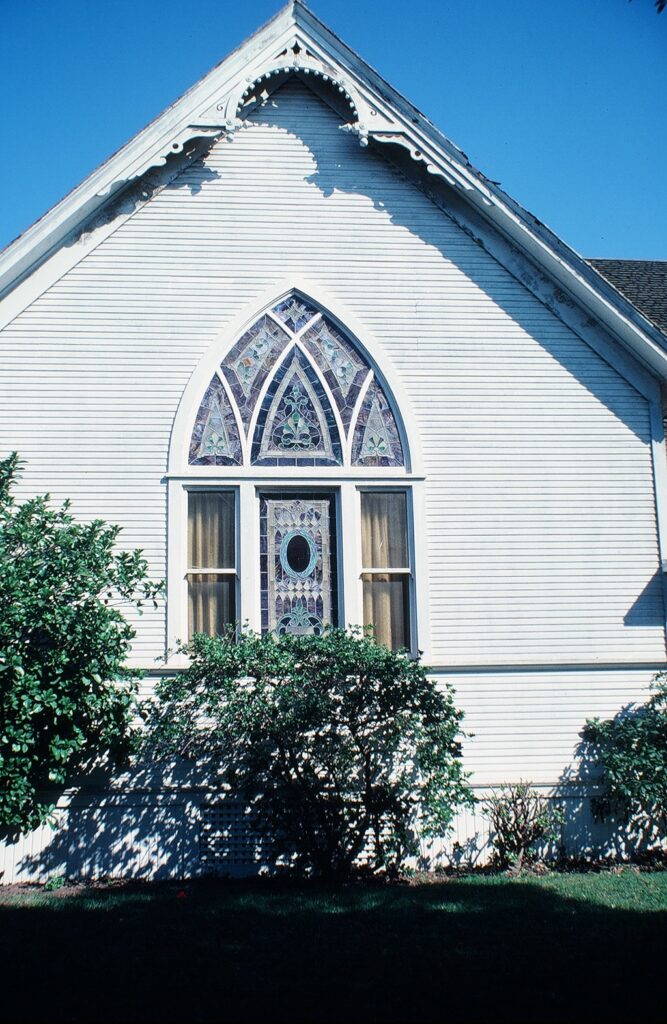 White church facade featuring a Gothic arched stained glass window above a door, surrounded by green shrubs under a clear blue sky; captured before relocation in 1982.