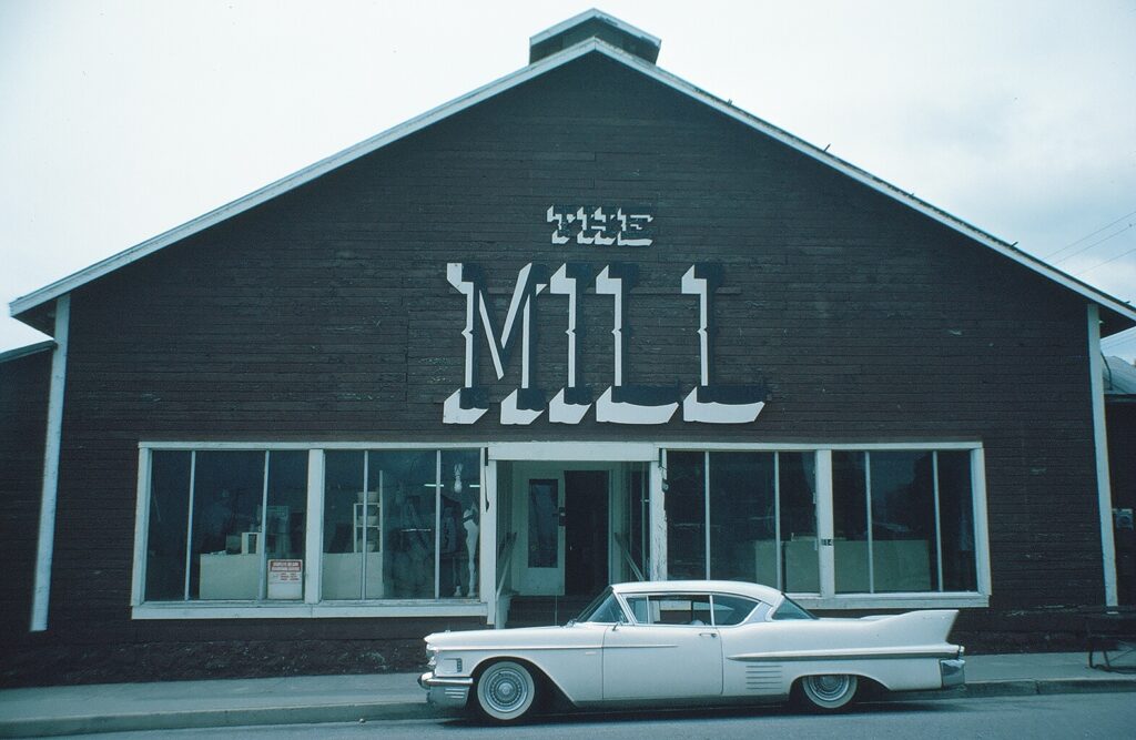 Dark brown wooden building with "The Mill" signage in large white letters, white vintage car parked in front, 1982.