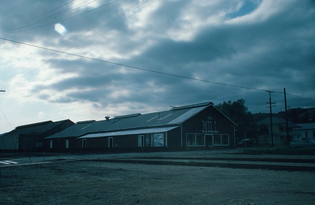 Dark industrial mill building labeled "MILL" stands beside railroad tracks under a cloudy sky in a 1982 photograph.