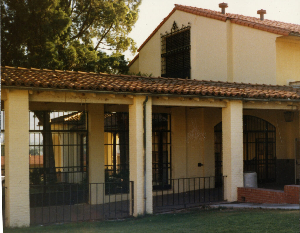 1985 photo of a beige building with a red-tiled roof, covered porch supported by columns, barred windows, and a grassy lawn.