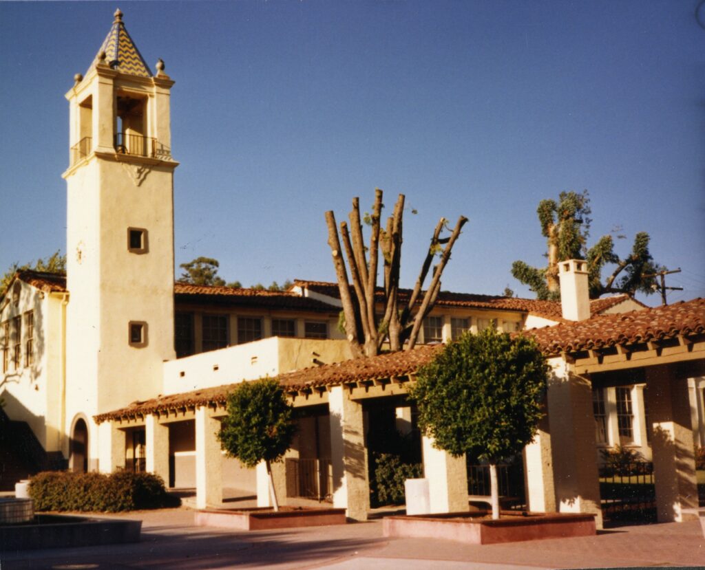 1985 photograph of a beige building with a bell tower featuring a blue and yellow tile roof, red-tiled main roof, and a covered walkway with columns, surrounded by trees under a clear blue sky.
