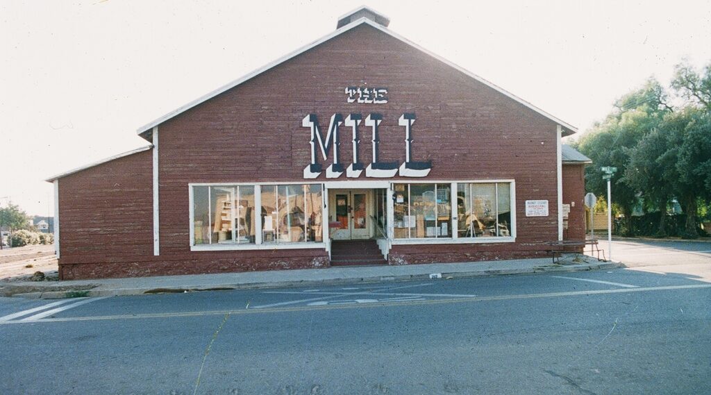 1985 photograph of The Mill, a red brick building with large "MILL" lettering above the entrance, display windows, and a parking lot prior to street reconfiguration.