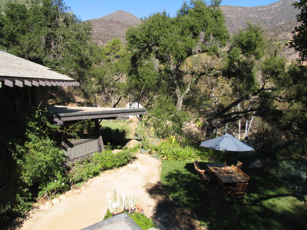 2007 exterior view of a house with a covered patio, outdoor dining table under a blue umbrella, garden, and mountain backdrop.