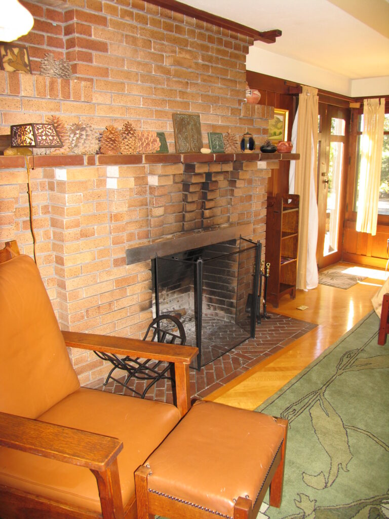 A 2007 interior living room features a brick fireplace with a mantel holding pinecones and decorative items, a brown leather chair with ottoman, and French doors with curtains allowing natural light. The space has wooden flooring and a green rug with tree patterns.