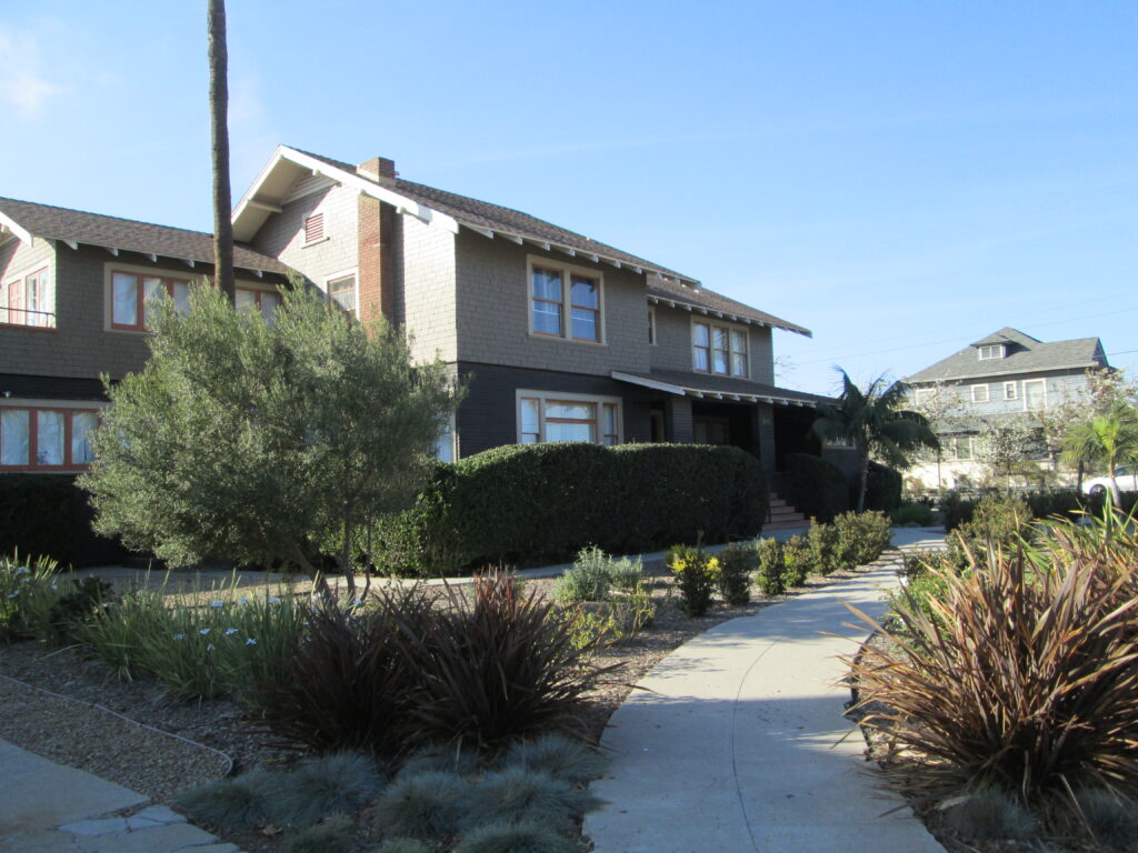 2018 photograph of a residential street with two-story houses, landscaped yards featuring shrubs and trees, and a paved sidewalk curving through the property under a clear blue sky.