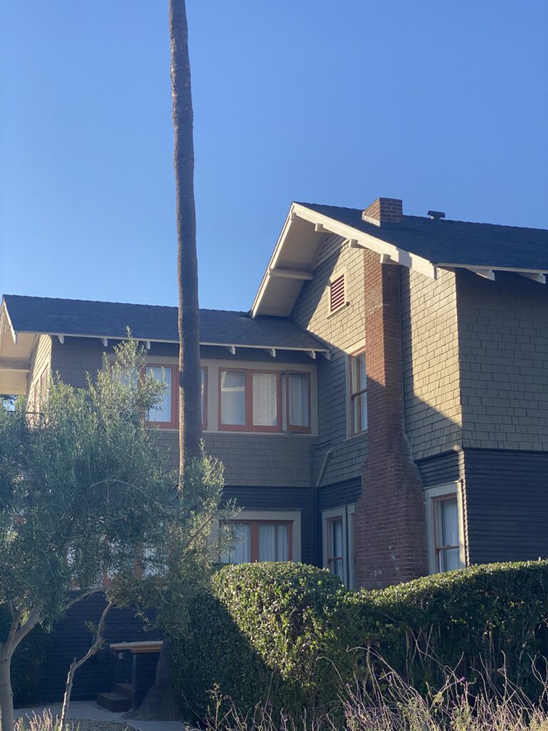 2021 photo of a house with brick and shingle siding, red-trimmed windows, and a palm tree against a clear blue sky.