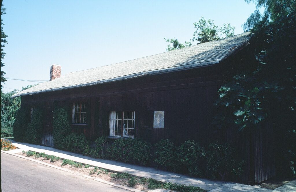 Dark wooden single-story barn with sloped roof, brick chimney, and greenery around windows, situated on a sidewalk near a street, labeled Artists' Barn circa 1980s.