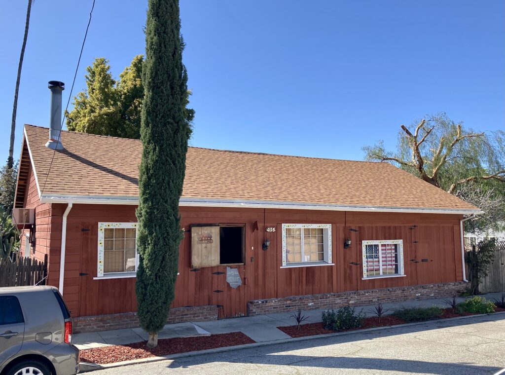 Red wooden Artists’ Barn with brown shingled roof, number 416 visible, decorative windows (sunflowers, American flag), tall tree in front, and a parked car.