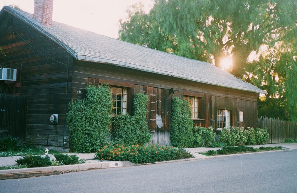 Dark wooden barn with shingled roof, number 416 visible, ivy-covered walls, windows, and orange flowers along the sidewalk, with trees and setting sun in background.