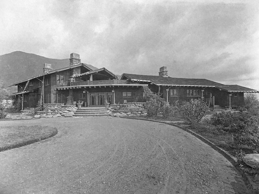 Black-and-white photograph of the Charles Millard Pratt House front elevation, featuring a wooden structure with stone chimneys and foundation, set against a mountain backdrop.