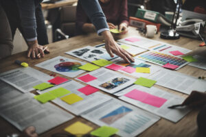 Team members collaborate around a table with documents, pink and green sticky notes, color swatches, and a document labeled "INFORMATION". Hands arrange materials during a design planning session.