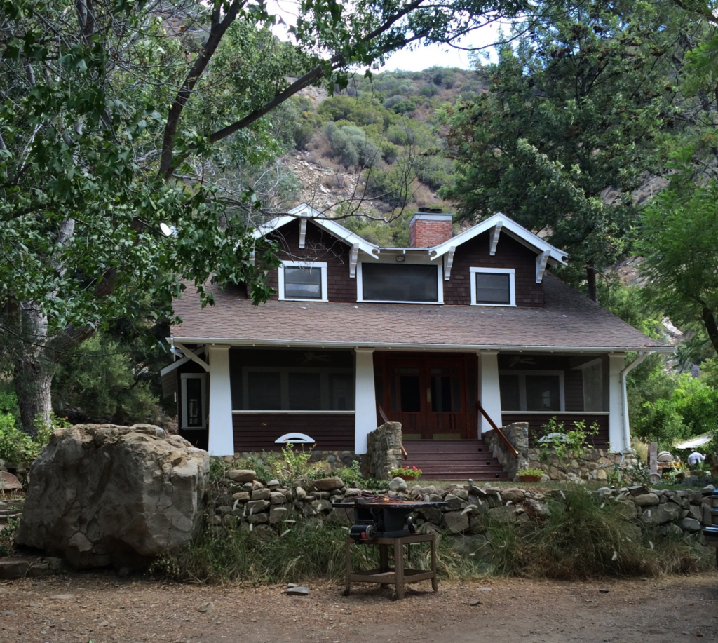 Brown shingle house with white trim and stone foundation, featuring a wooden front porch with stairs, surrounded by trees and a rocky hillside.