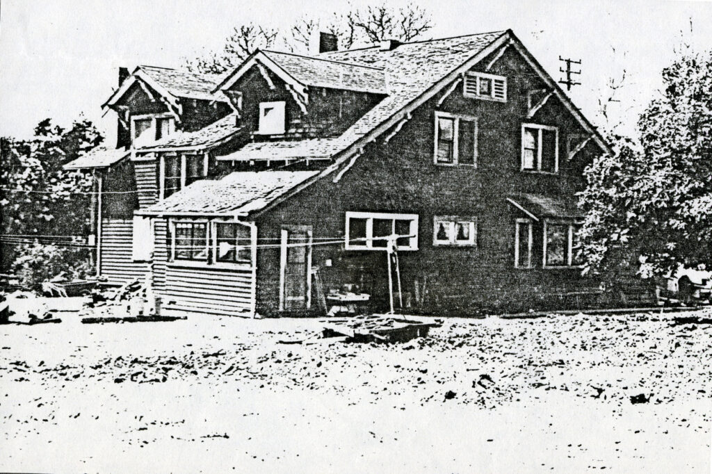 Black-and-white photograph of the Dent Ranch House, circa 1980s, showing a two-story structure with a covered porch, multiple windows, and a yard with scattered debris.