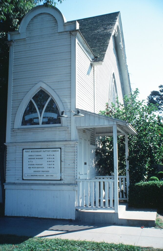 White wooden First Missionary Baptist Church with Gothic windows and a sign listing Sunday school and worship times; photographed before relocation in 1982.
