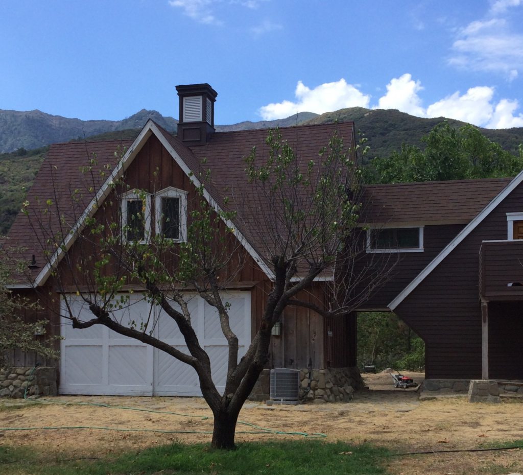 Brown wooden house with white garage doors and dormer windows, featuring a tree in the foreground and mountains in the background.