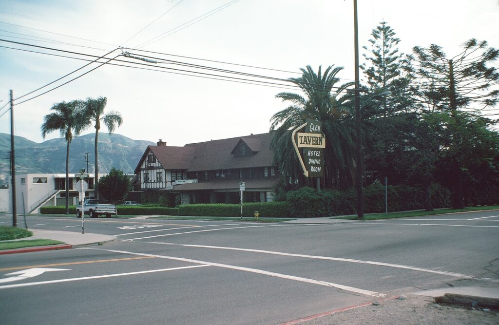 1982 photograph of Glen Tavern Hotel with its sign advertising "Hotel Dining Room" and Tudor-style architecture, set at a street intersection with palm trees and mountains.