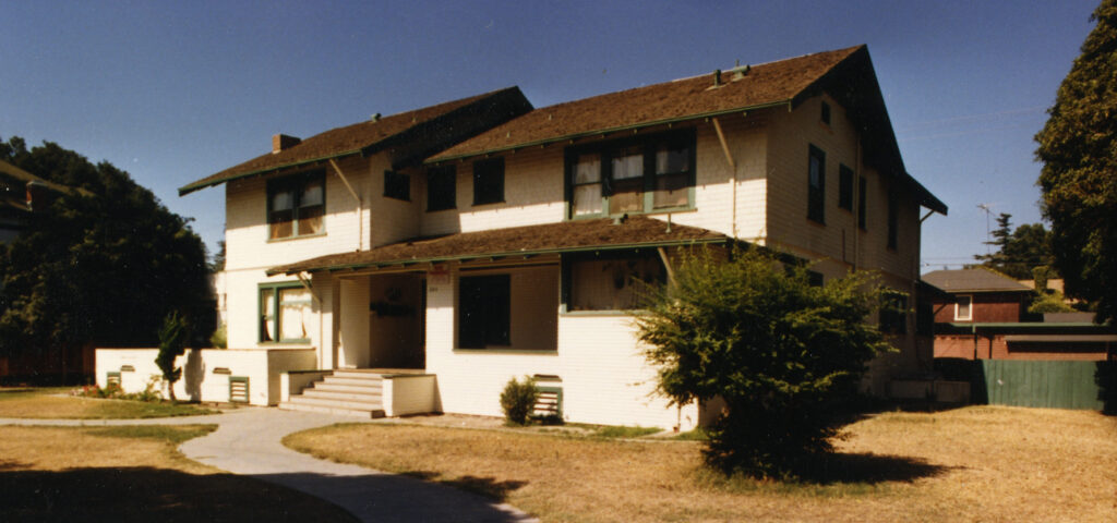 Two-story white house with green trim and brown shingles, featuring a front porch with steps, surrounded by trees and a lawn under a clear blue sky. Henry Levy House, circa 1980s.