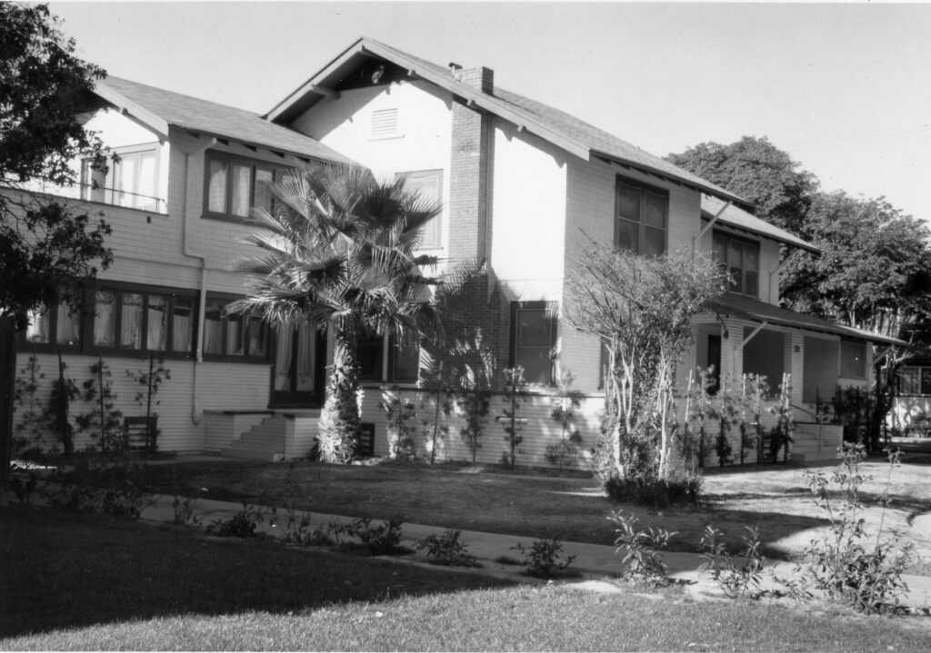 Two-story house with gabled roof, brick chimney, and palm tree in front, surrounded by landscaping and trees; circa 1980s.
