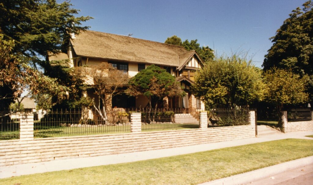 Two-story Henry Levy House with thatched roof, surrounded by trees and a brick fence, circa 1980s.