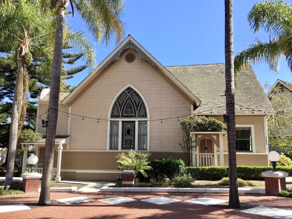 A beige Victorian-style church with a gabled roof and stained glass window at Heritage Square, featuring palm trees, a brick walkway with white squares, and the address number 731 visible on the facade.