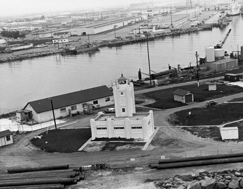Black-and-white historical photo of a harbor with a central lighthouse, industrial buildings, boats, and a road with a car. The scene depicts waterfront infrastructure and watercraft.