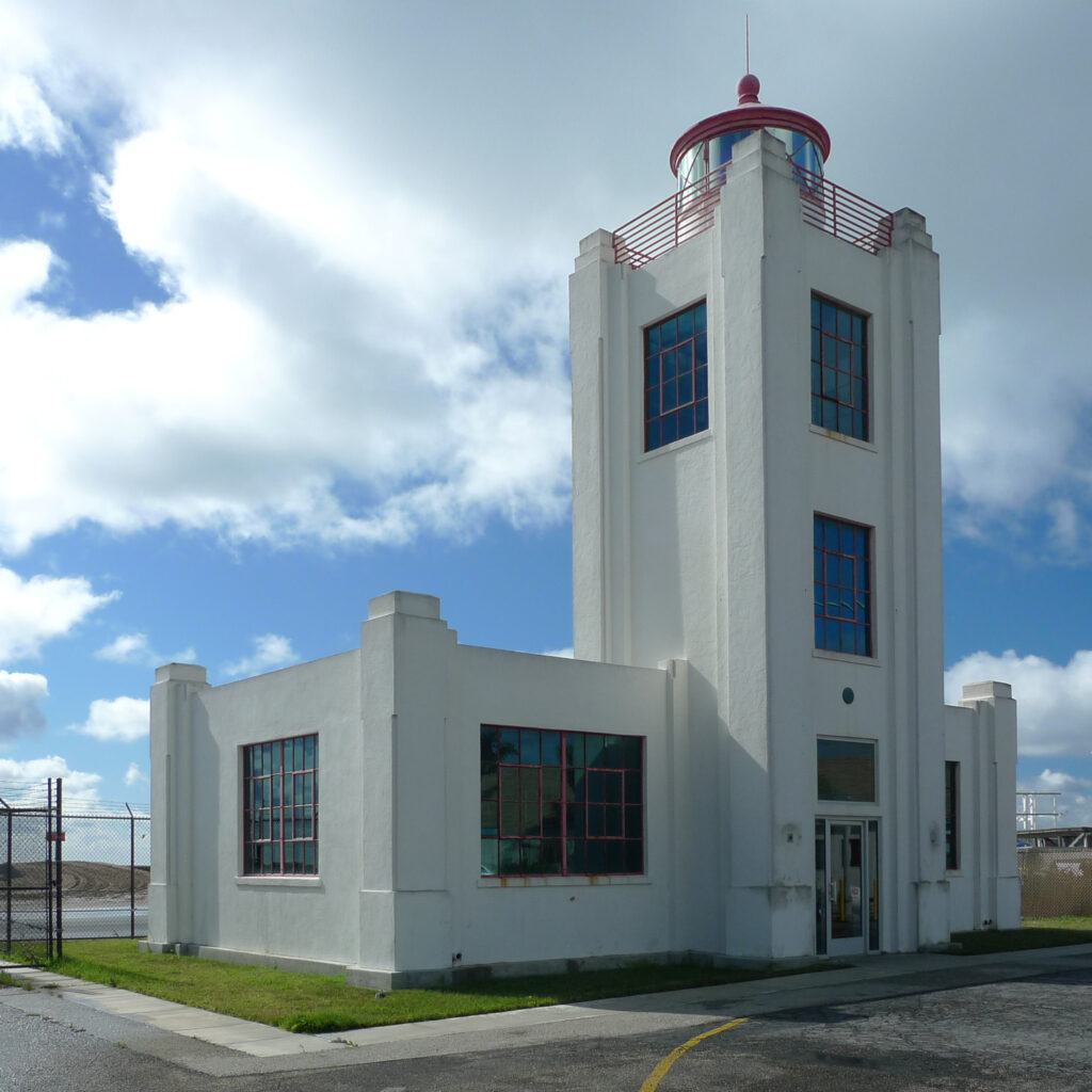 White Hueneme Lighthouse with red accents, featuring a tower with a red dome and railing, large windows with red frames, and a main building with a door, set against a partly cloudy sky.