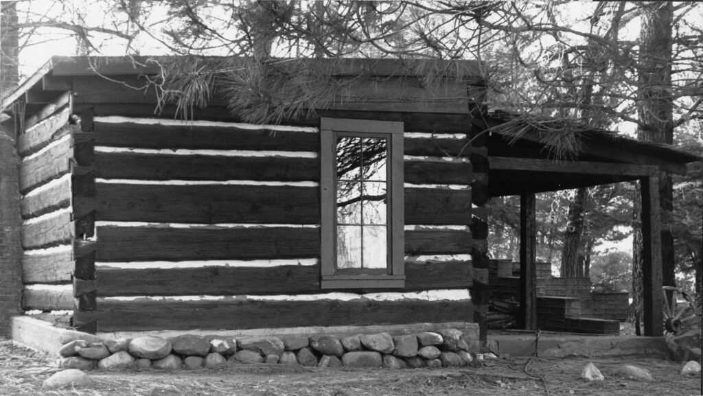 Log cabin with horizontal logs, white chinking, window, stone foundation, and covered porch surrounded by trees. Lathrop Camp, circa 1980s.