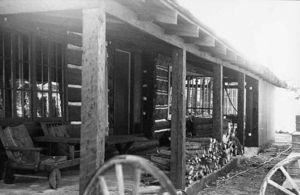 Black-and-white photograph of Lathrop Camp circa 1980s, showing a log cabin porch with wooden chairs, stacked firewood, and wagon wheels in the foreground.